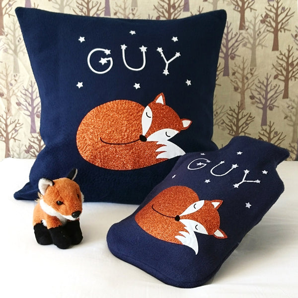 A navy fleece hot water bottle cover and matching cushion featuring a sleeping fox with glowing star constellations spelling out a child's name.