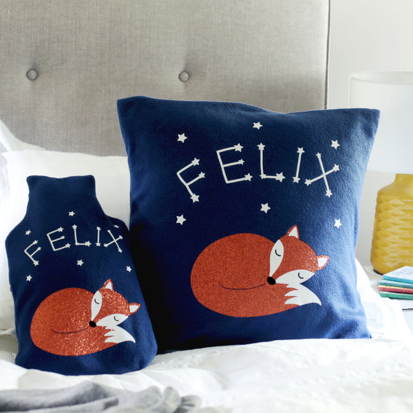 A cute and comforting hot water bottle cover with matching cushion in navy fleece, with a sleeping fox and glow-in-the-dark star constellations forming a child's name.
