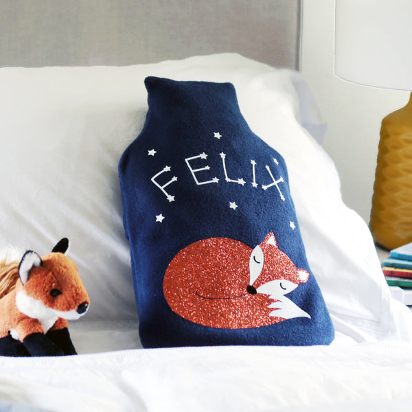 A snuggly navy fleece hot water bottle cover with a whimsical sleeping fox design and personalized glow-in-the-dark star constellation spelling out a child's name.