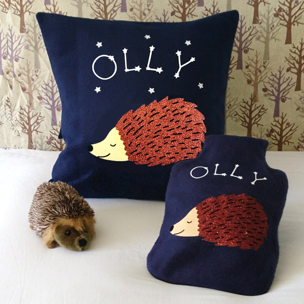A comforting hot water bottle cover and matching cushion  in navy fleece, with a sleeping hedgehog and glow-in-the-dark star constellations forming a child's name.