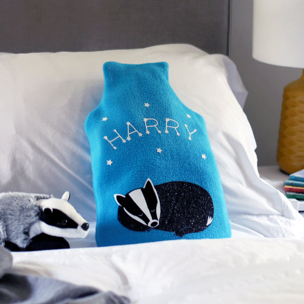 A cosy hot water bottle cover featuring a sleeping baby badger surrounded by constellations and stars. The turquoise fleece is super-soft and perfect for chilly evenings.