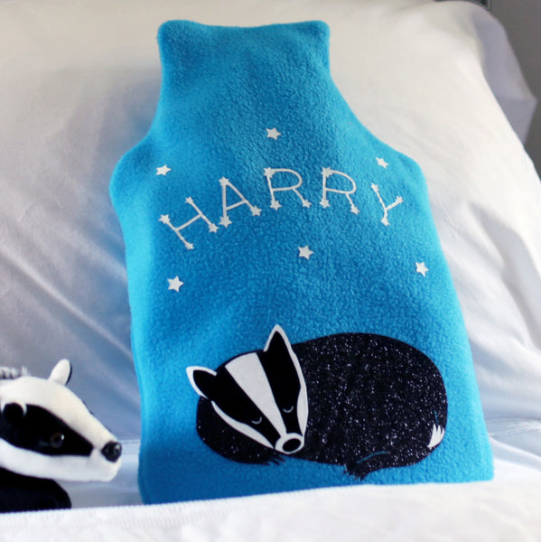 A soft and snuggly hot water bottle cover with a whimsical baby badger design, featuring constellations and stars in the background. The cover is perfect for keeping warm on cool nights.