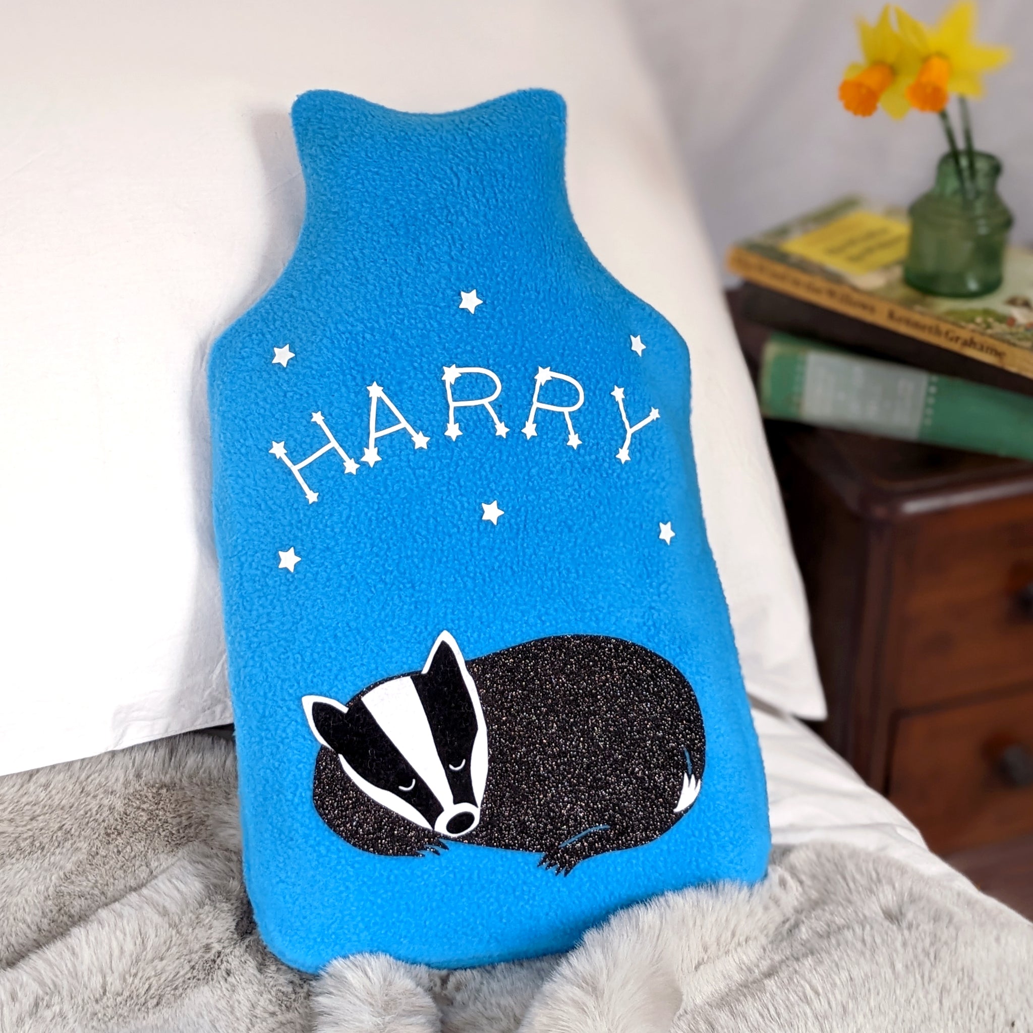 A photo of a hot water bottle cover in turquoise fleece with a cute baby badger design. The badger is sleeping soundly under a starry night sky filled with constellations and twinkling stars.